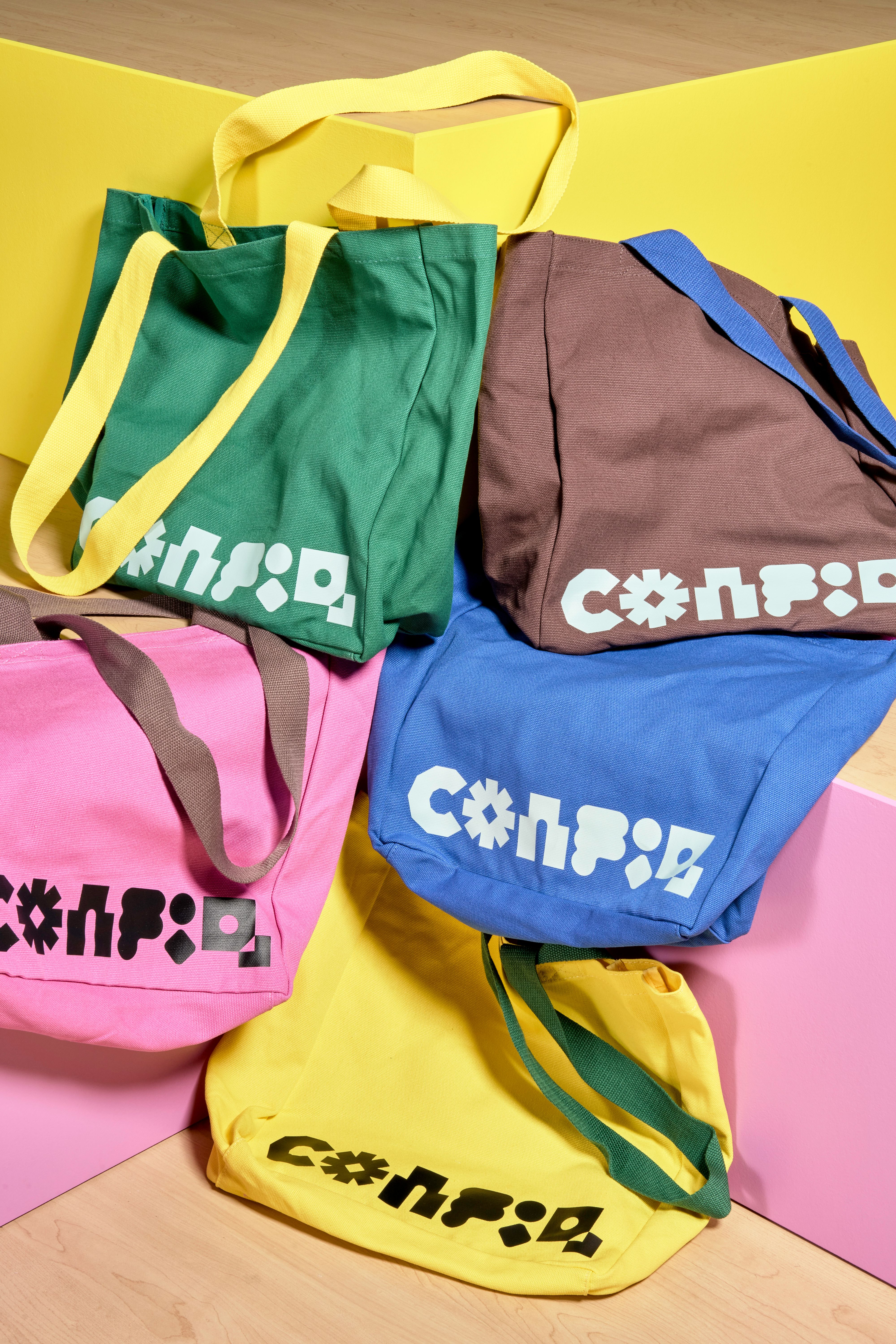 There are five totebags loosely arranged on a pink and yellow stepped structure. Each bag is a different color—green, brown, blue, yellow, and pink—with a Config logo on the bottom edge. 