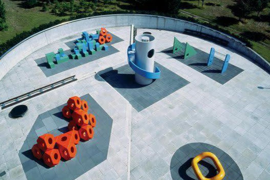 An outdoor playground featuring abstract, colorful climbing structures and play equipment arranged on a large tiled surface. Bright orange geometric shapes resembling clusters of triangular blocks sit on one side, while a tall cylindrical structure with a blue spiral slide stands at the center. Other green, blue, and yellow structures, including a triangular climbing wall and a yellow ring-shaped object, are spread across the area. The playground is surrounded by a low circular wall, with grass and trees visible beyond.