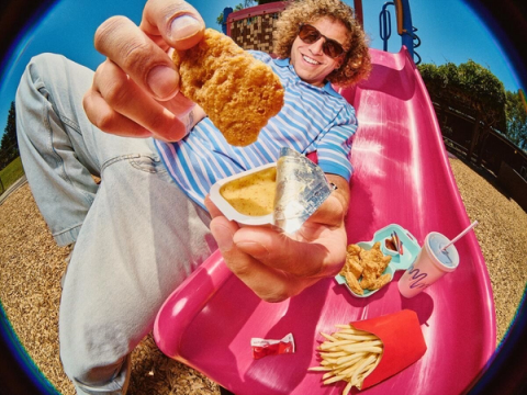 Smiling man in sunglasses poses next to a pink playground slide, holding a chicken nugget.
