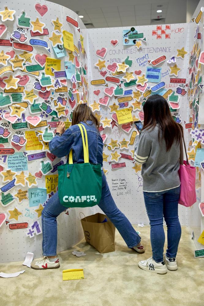 Two people stand in front of a white wall that is covered with different colored stickies. Each stickie has a hand-drawn message. The person on the left has their legs outstretched, helping them balance while writing a message on the middle of the wall.