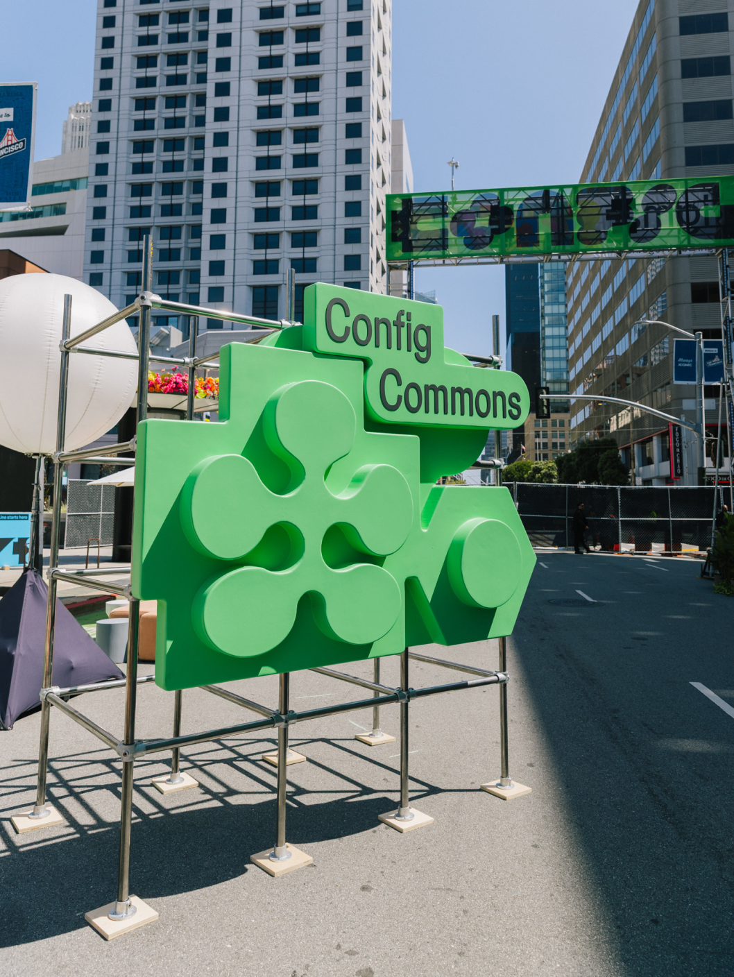 Bright green “Config Commons” sculpture stands on scaffolding in street surrounded by tall downtown buildings.