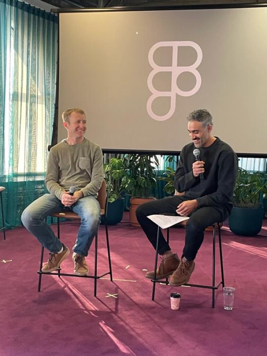 A photograph of two men sitting on stools in front of a screen with the Figma logo. They're smiling and holding microphones.