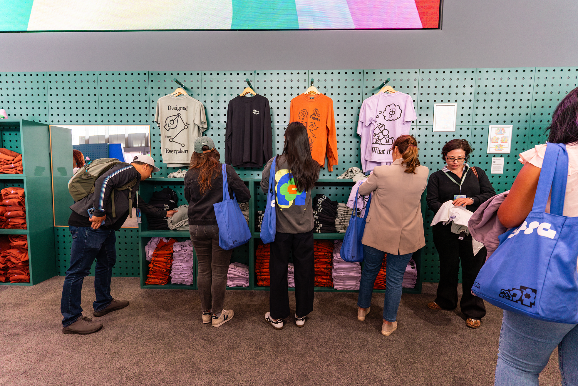 Several attendees select Figma-themed shirts from a green pegboard display wall, examining designs and folding clothes while carrying blue tote bags.