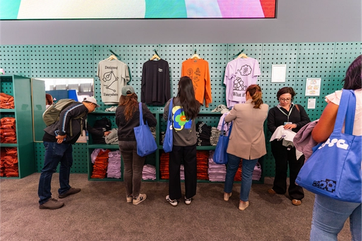 Several attendees select Figma-themed shirts from a green pegboard display wall, examining designs and folding clothes while carrying blue tote bags.