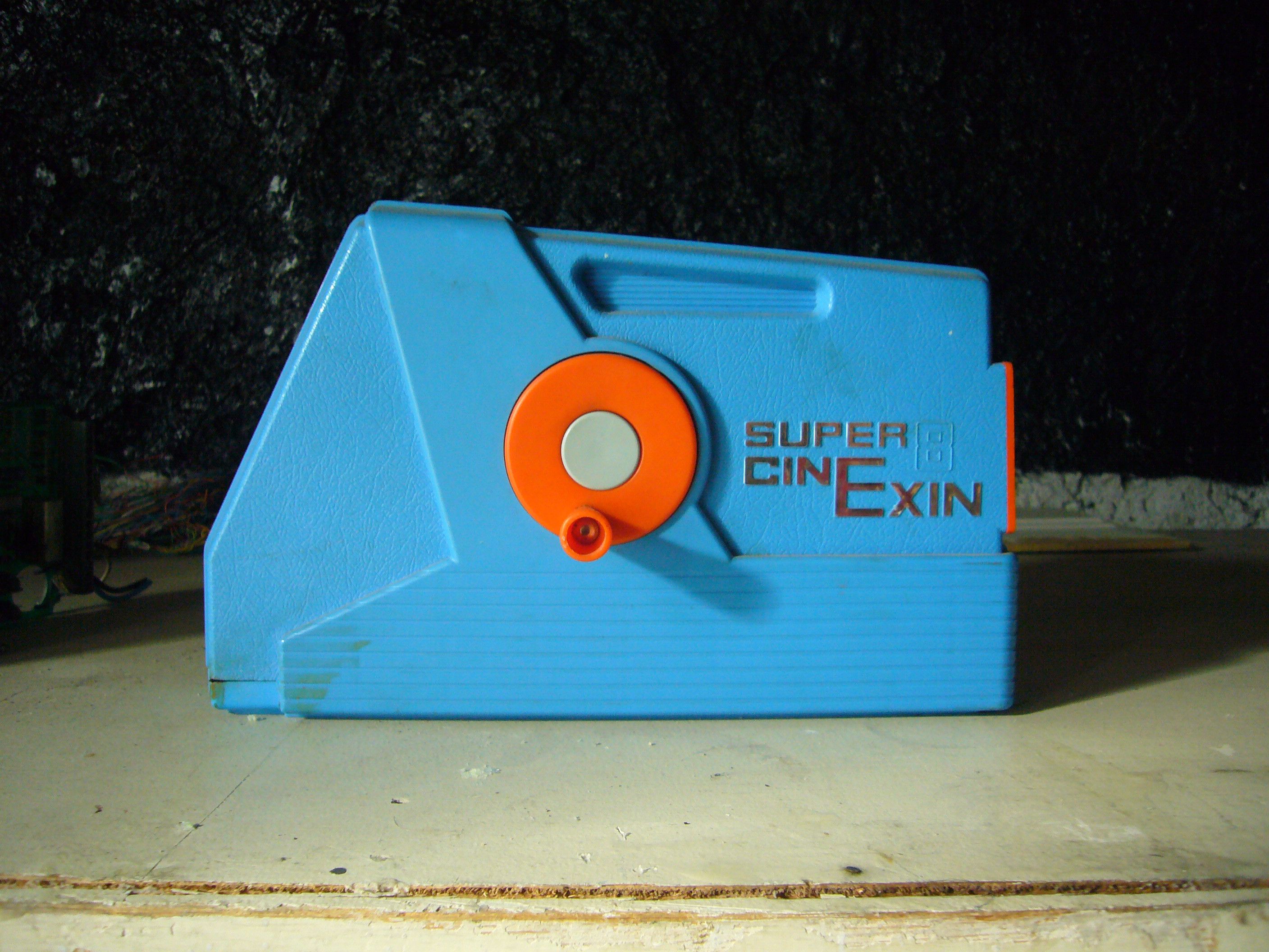A vintage blue Super CinExin 8mm film projector with an orange knob, placed on a light-colored surface against a dark background.