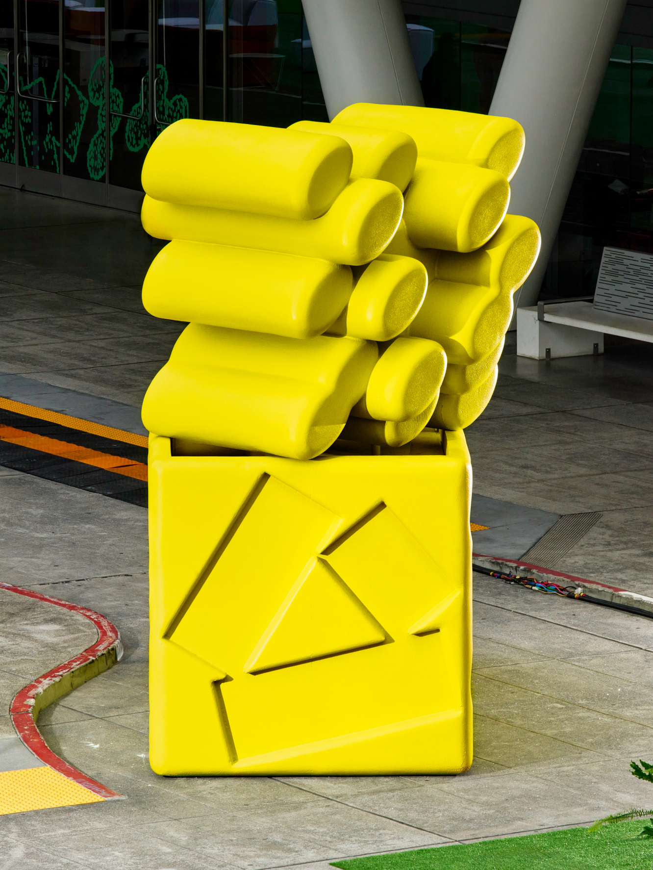A large yellow sculpture combining a cube base and a cluster of overlapping cylindrical forms