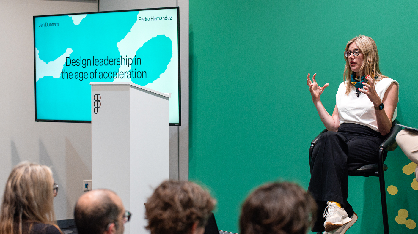 Ein Redner gestikuliert von einem Hocker neben einer Folie mit dem Titel „Design Leadership im Zeitalter der Beschleunigung“ aus und spricht zu den sitzenden Zuhörer*innen.