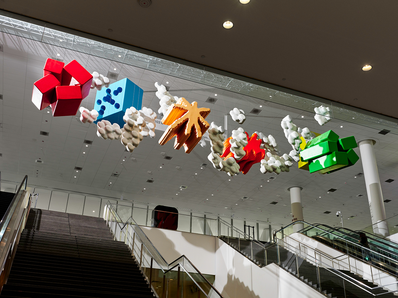 A row of colorful abstract sculptures—red cubes, a blue dice-like block, star shapes, a red splash form, and green blocks—hangs from the ceiling above escalators in a convention center atrium.
