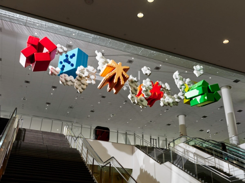 A row of colorful abstract sculptures—red cubes, a blue dice-like block, star shapes, a red splash form, and green blocks—hangs from the ceiling above escalators in a convention center atrium.
