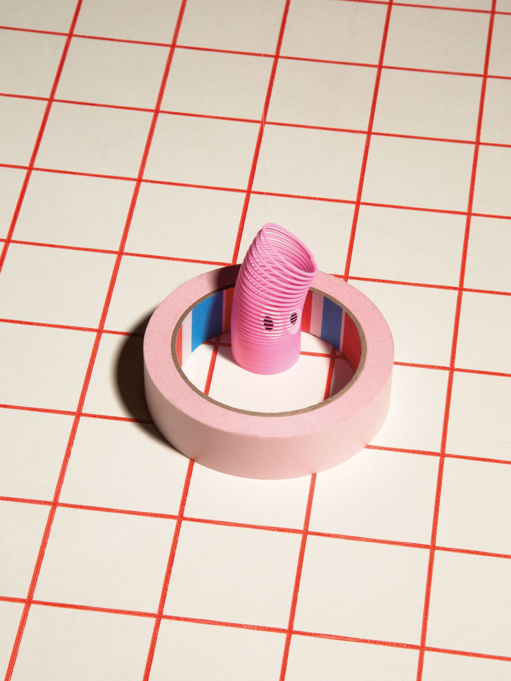 Pink toy spring standing inside a roll of pink tape on a red grid surface.