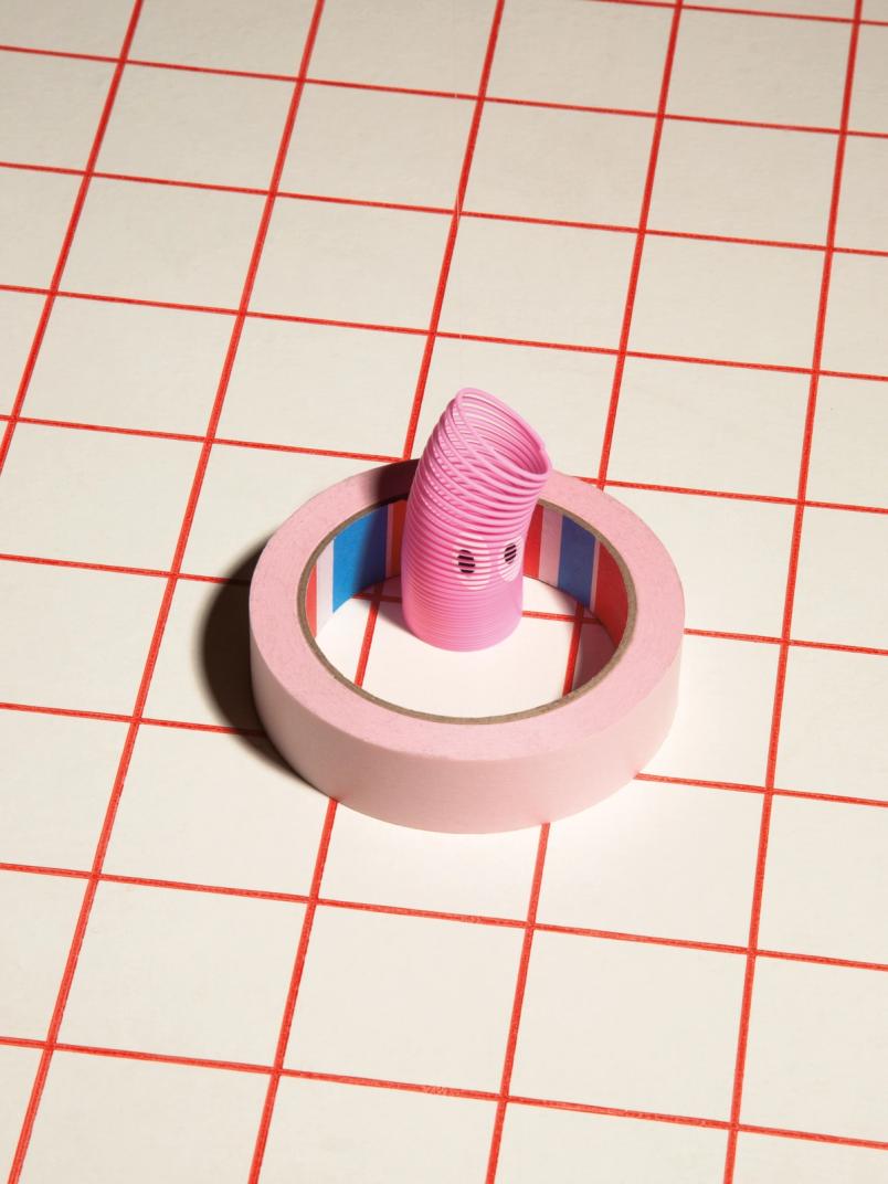 Pink toy spring standing inside a roll of pink tape on a red grid surface.