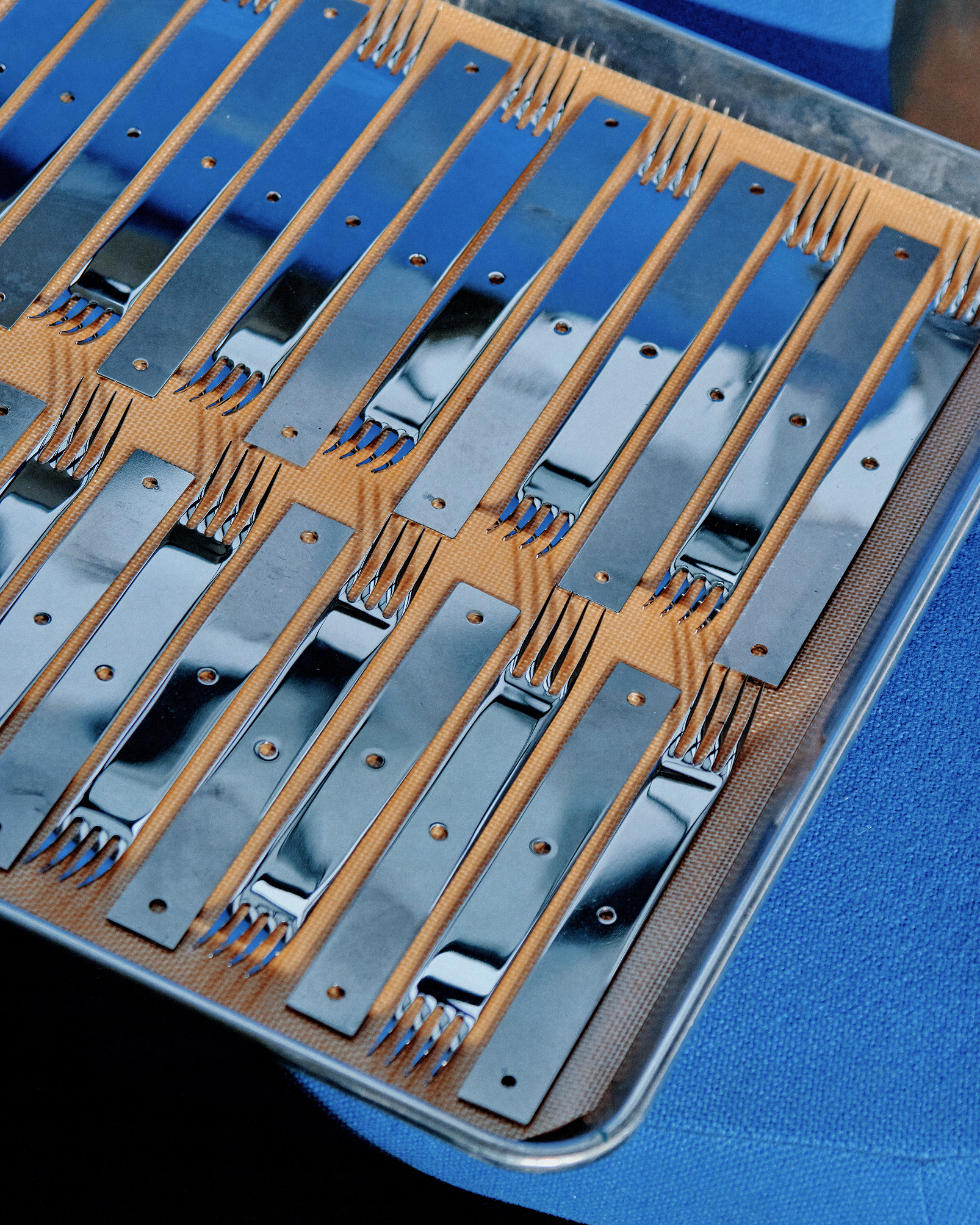 Tray holds neatly arranged, reflective fork pieces lined in rows on textured tan surface.