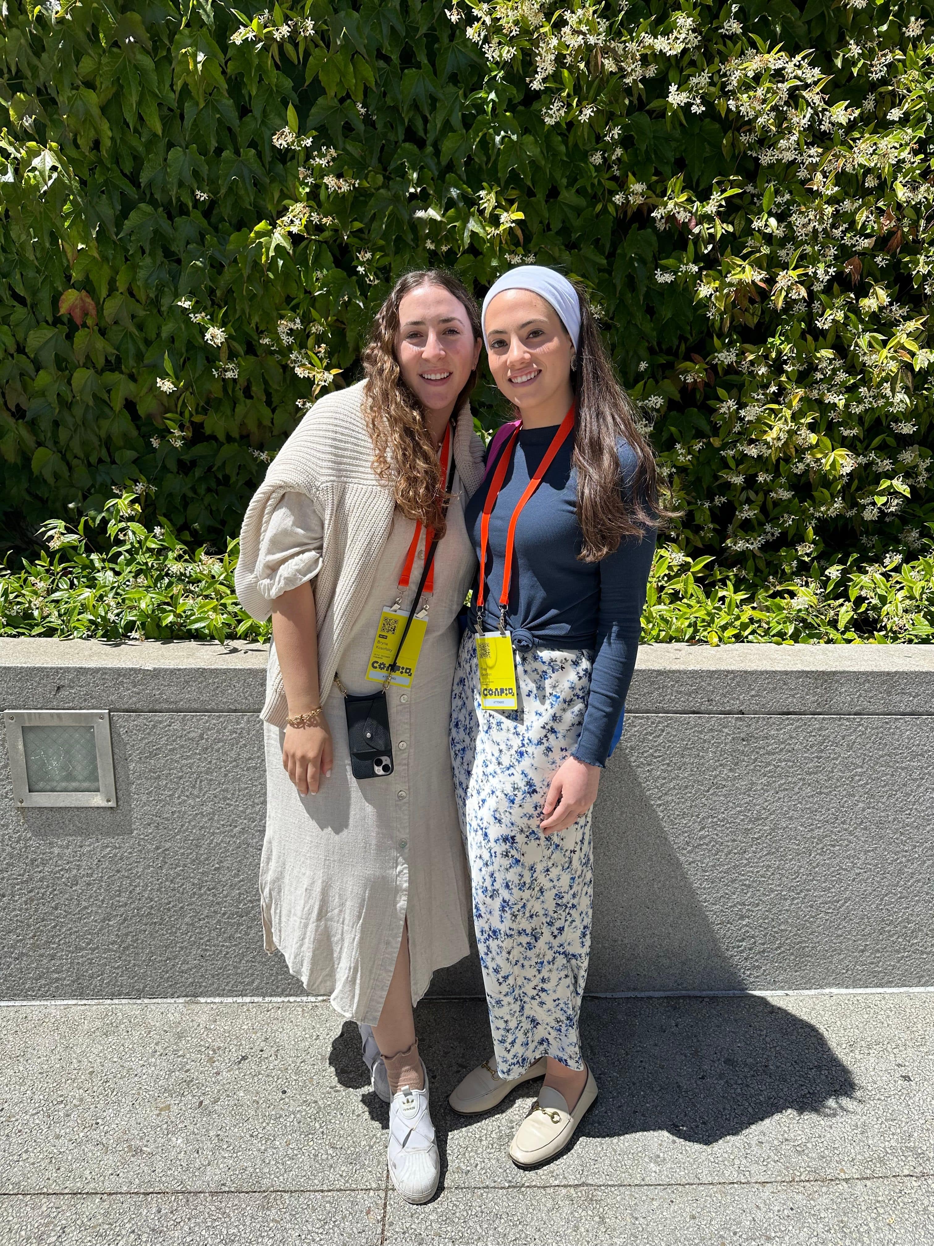 A woman wears a beige sweater draped over a beige maxi dress, and another wears a blue long sleeve tee and a floral maxi skirt.
