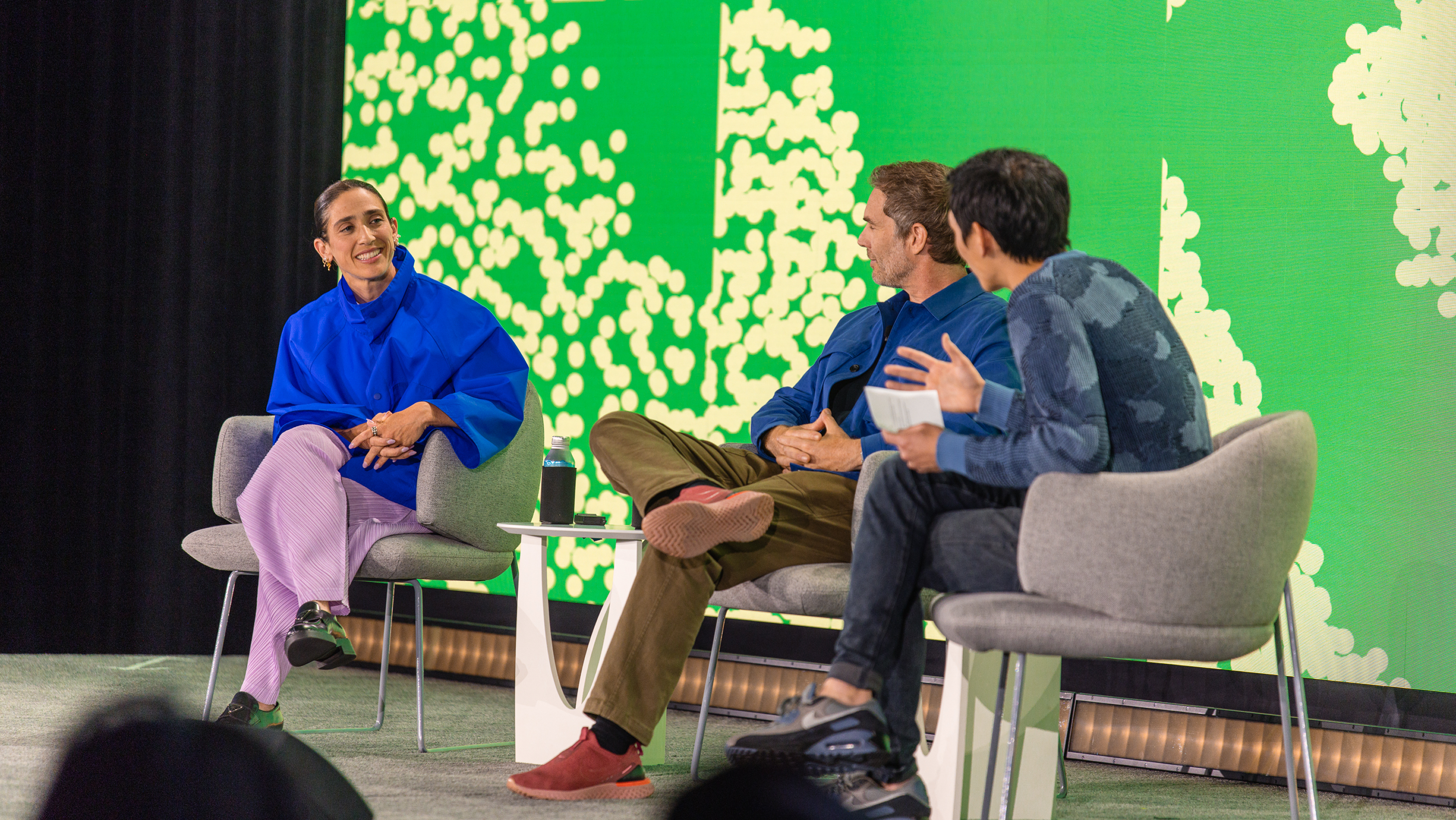 Rachel Been, Charlie Sutton, and Yuhki Yamashita sit in gray armchairs on a stage in front of a bright green patterned screen, mid-conversation.