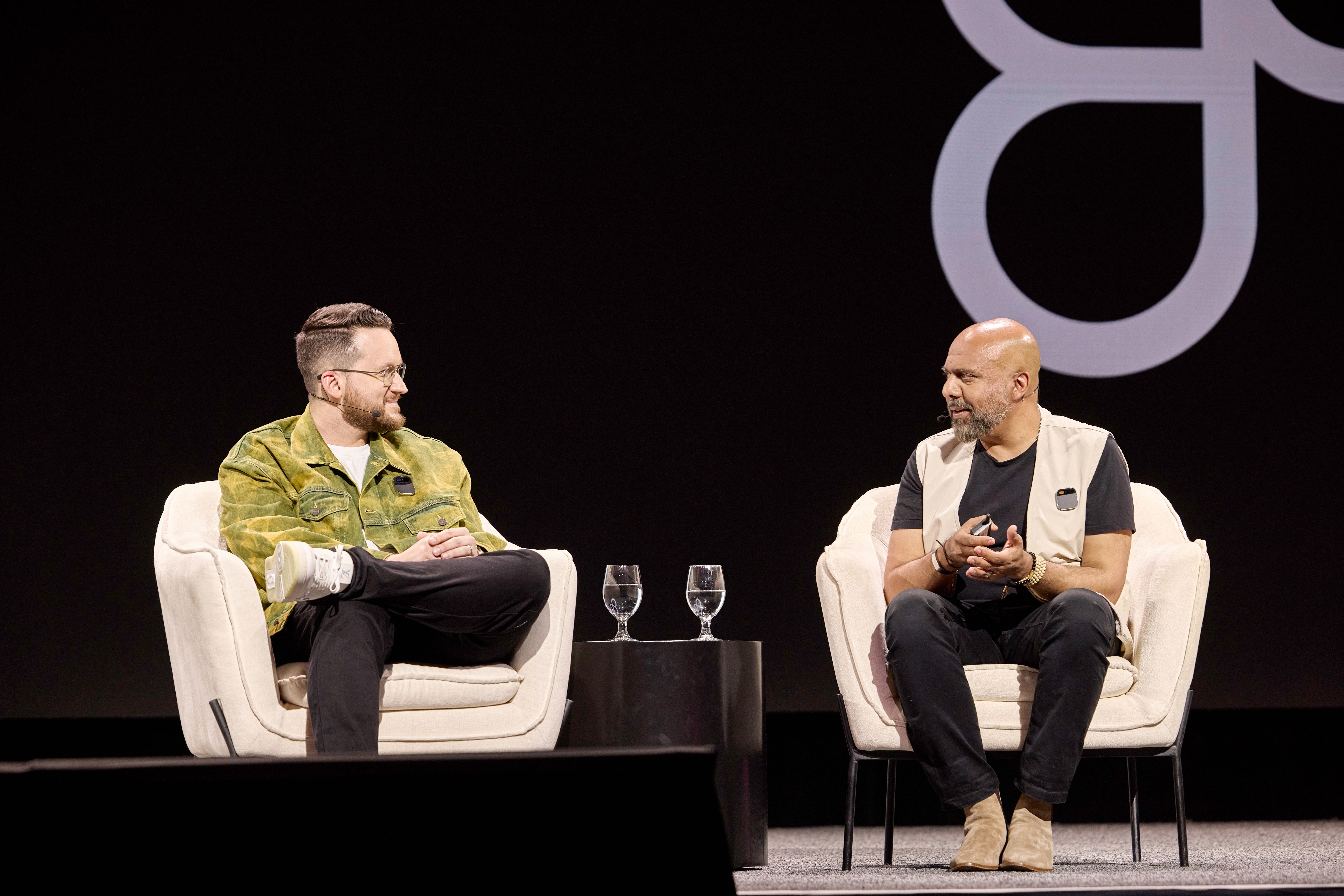 Two speakers sit in modern armchairs on stage, engaged in conversation. A small table with two glasses of water is placed between them. The background is dark with the Figma logo subtly displayed.