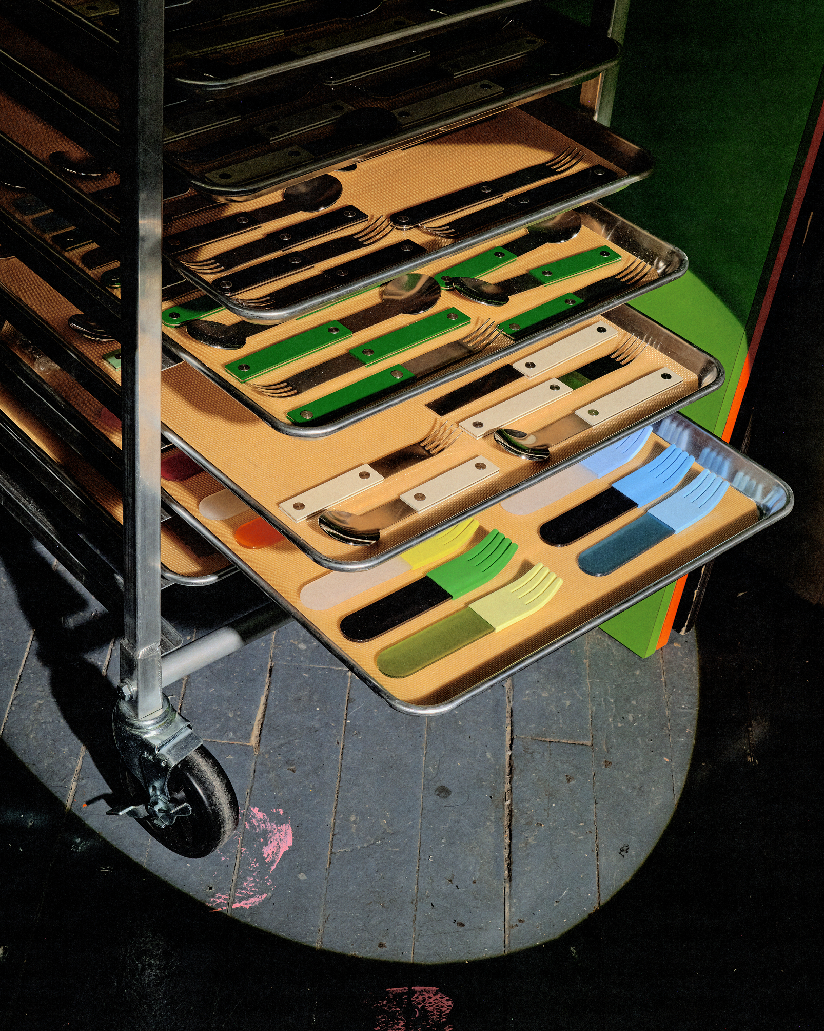 Metal cart holding trays of colorful handled utensils arranged neatly under dramatic lighting on worn floor.