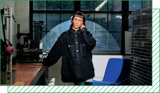 Photograph of a woman in an oversize jacket in an industrial studio, illuminated by a bright spotlight.