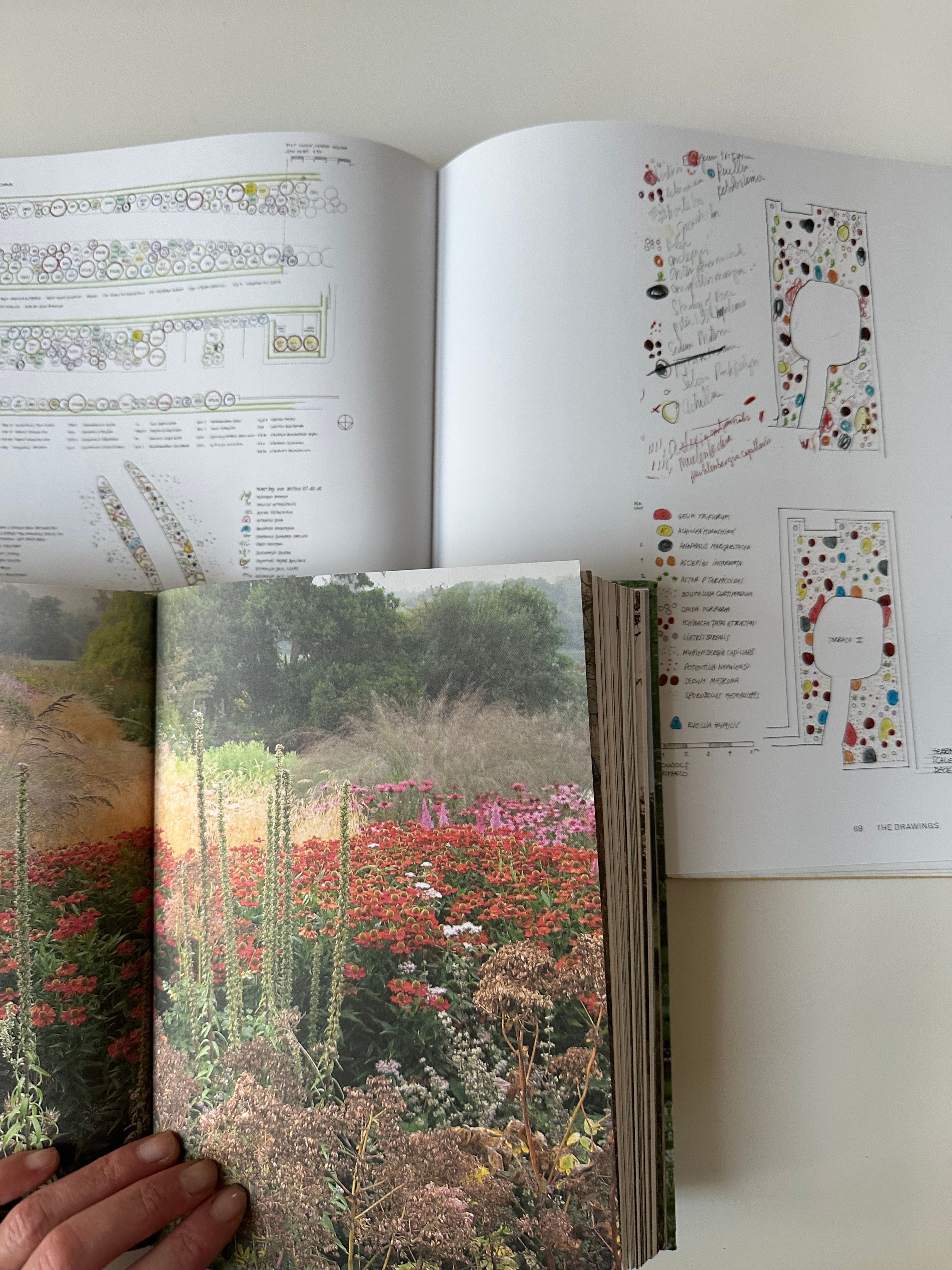 Two open books displayed on a white surface. The top book shows detailed garden or landscape design plans with handwritten notes and color-coded diagrams. The bottom book features a full-page photograph of a vibrant flower garden with a variety of colorful blooms and tall grasses. A person's hand is holding the bottom book open.