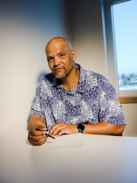 A portrait of Ron Scott, a Black man in a blue Hawaiian shirt, sitting in front of a window