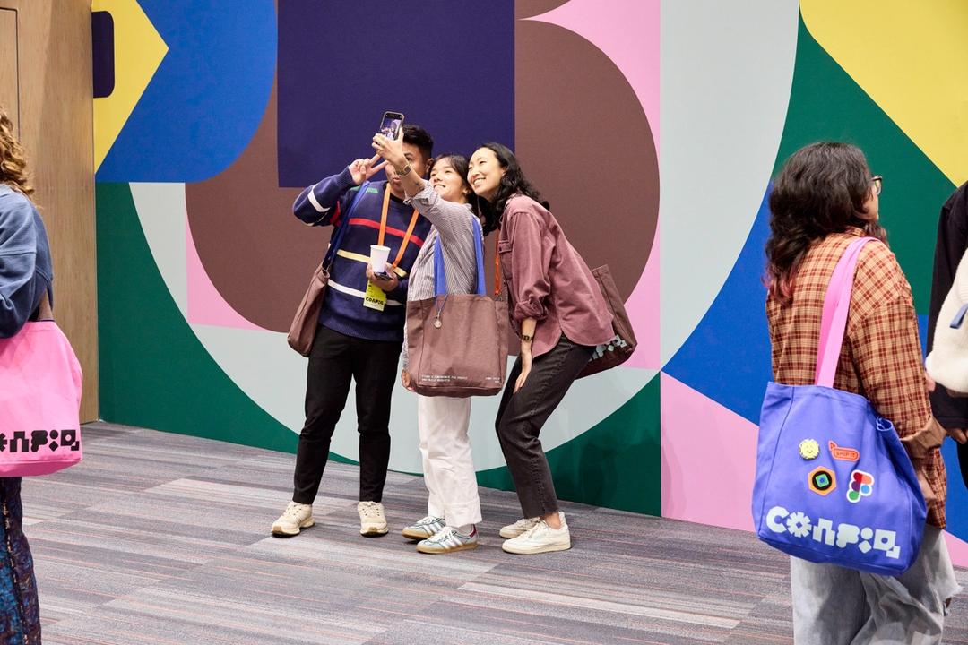 A group of three attendees posing for a selfie at a colorful conference backdrop. They are smiling, holding tote bags with conference branding, and wearing event lanyards. The wall behind them features a geometric design with bright colors, including pink, brown, green, and blue shapes.
