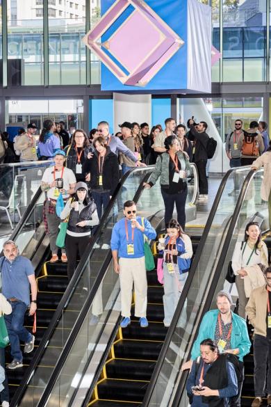 A busy scene with people using escalators inside a modern building, some wearing conference lanyards and engaging with one another.