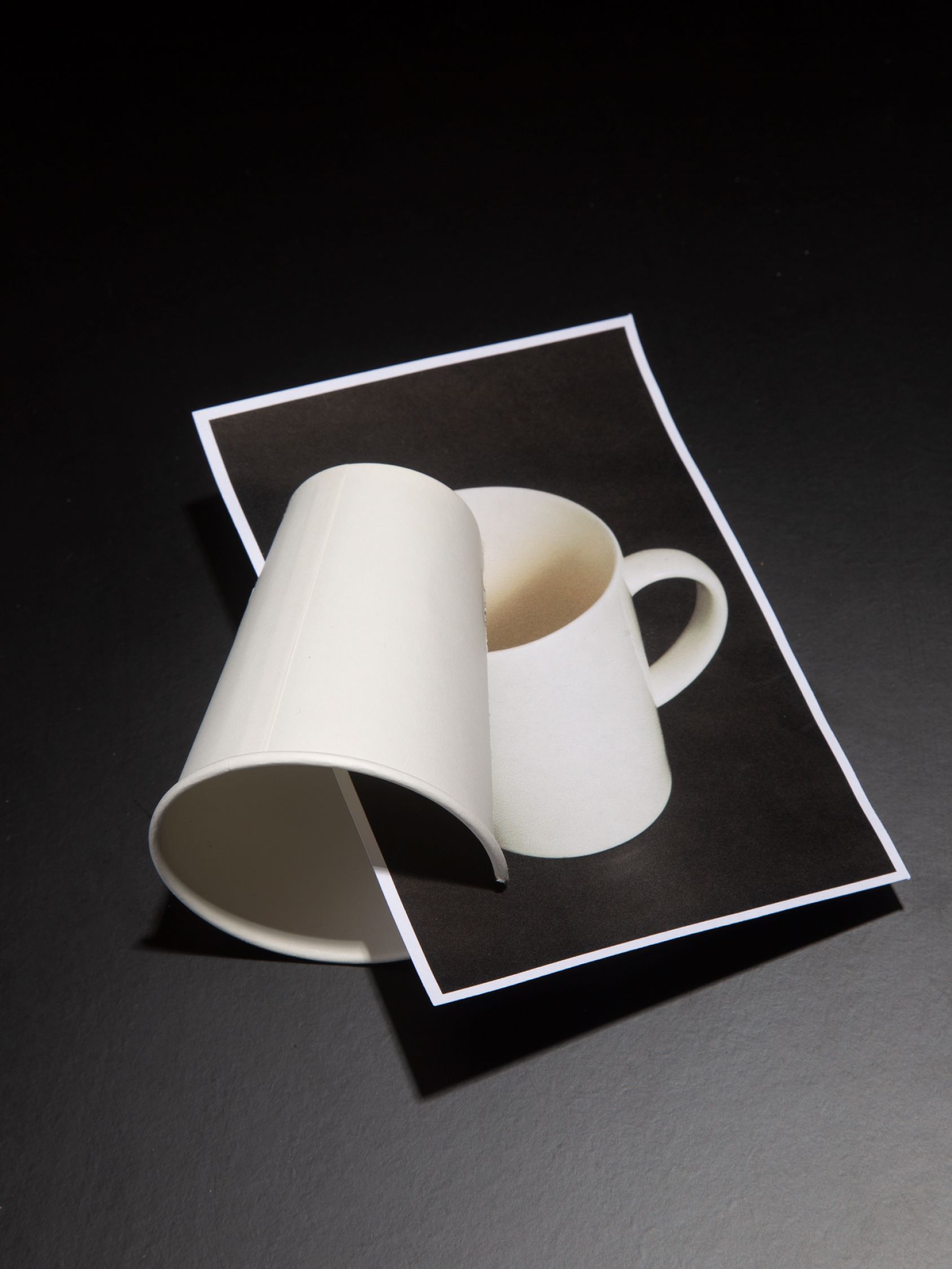 Curved white cup placed over a printed photo of a matching coffee mug, creating a visual illusion.