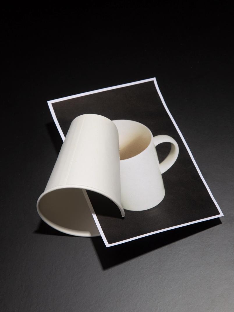 Curved white cup placed over a printed photo of a matching coffee mug, creating a visual illusion.