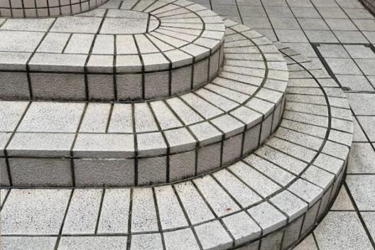 A tiled outdoor staircase with curved steps, made up of rectangular light gray tiles with dark grout lines. The steps are rounded and arranged in a semi-circular fashion, leading to a higher platform. A cylindrical tiled column is visible on the left, with small green plants growing near its base. The design emphasizes clean lines and geometric symmetry.