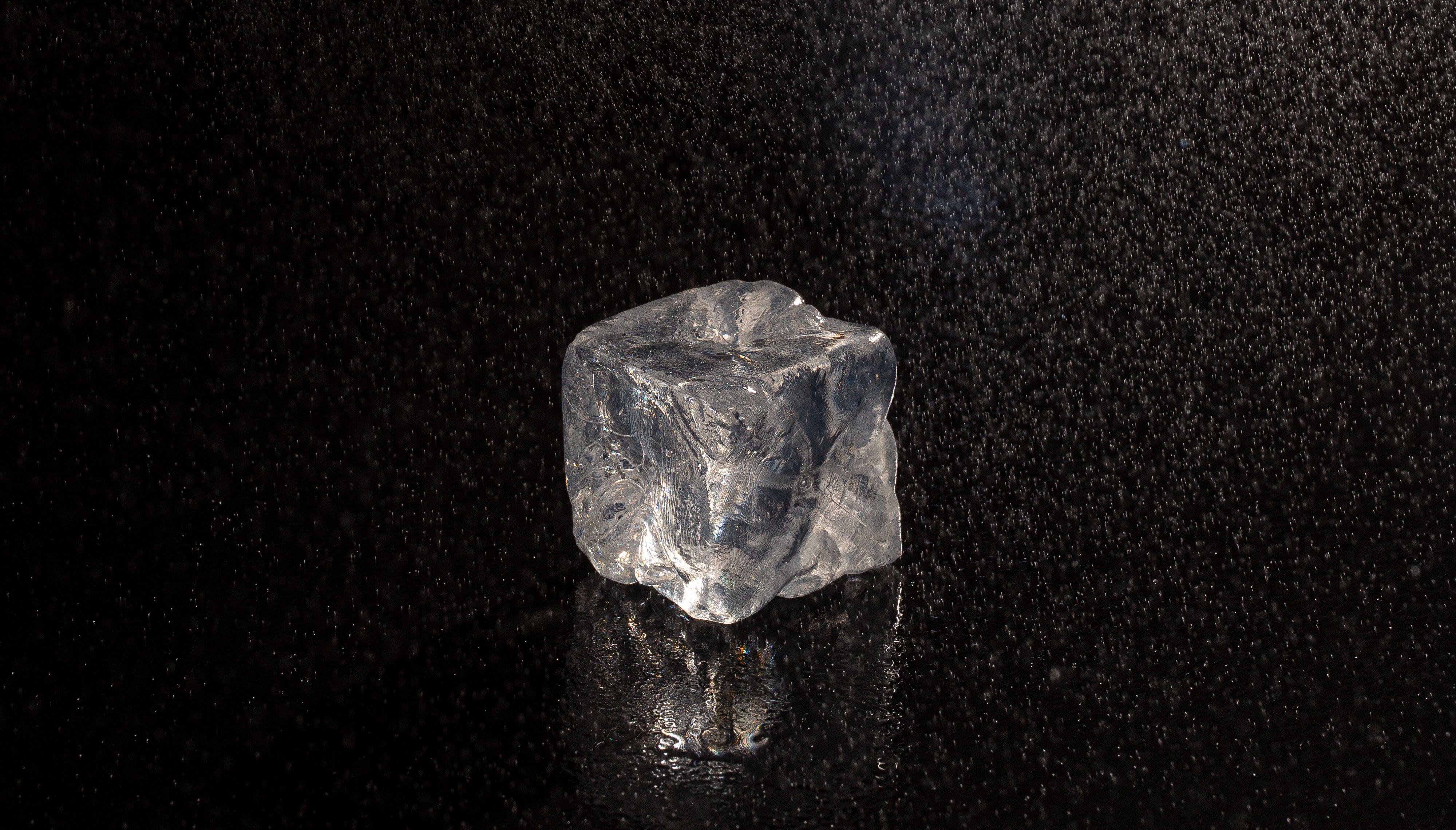 Transparent ice cube on a wet black surface with water droplets falling around it.