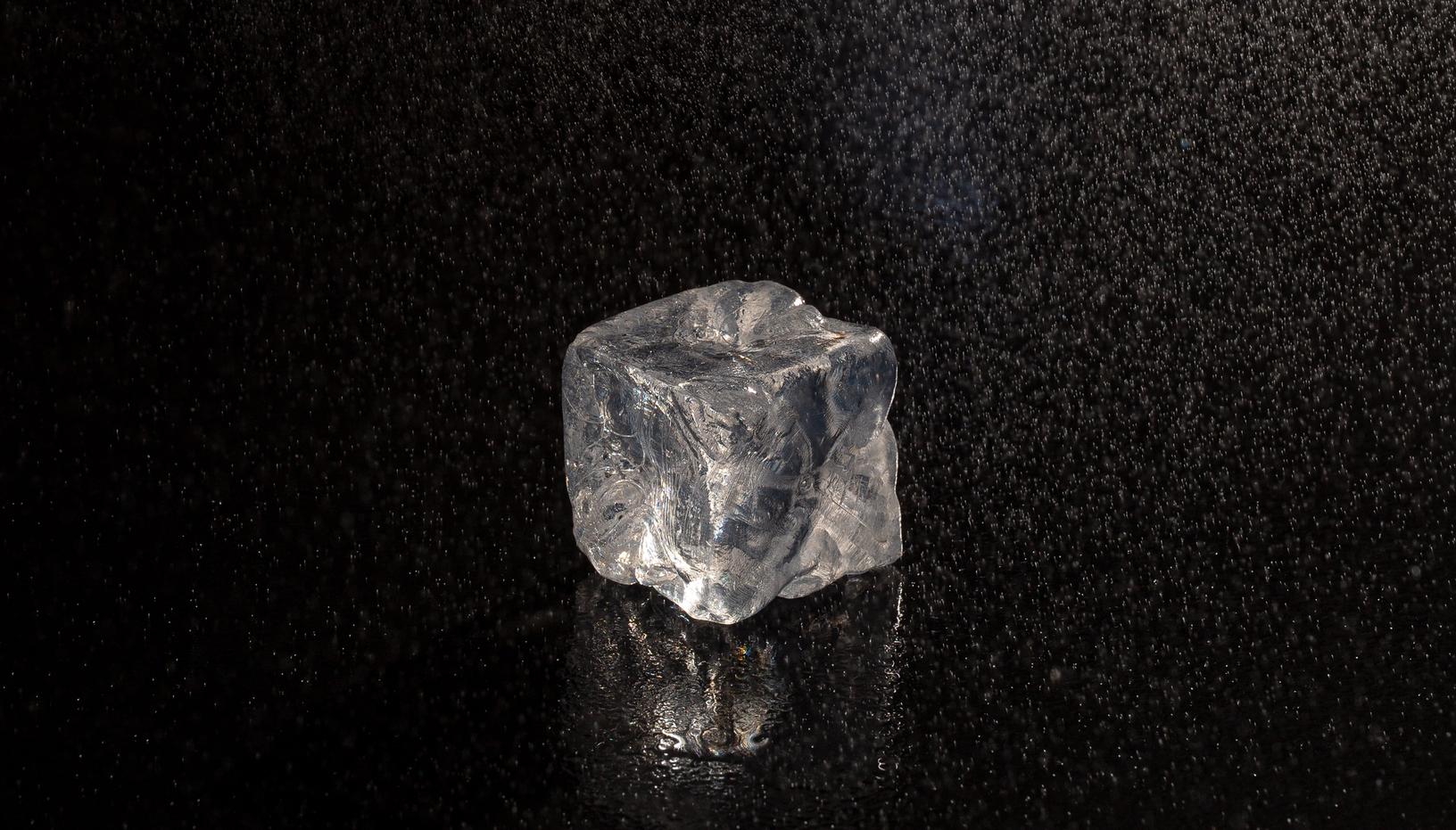 Transparent ice cube on a wet black surface with water droplets falling around it.