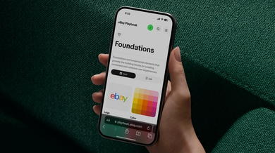 A hand holding a smartphone displaying the "eBay Playbook" design system on a dark green fabric background. The screen shows the "Foundations" section with options for grid or list views and colorful tiles representing logo and color guidelines.