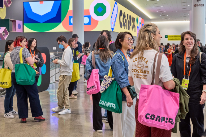 In the lobby of a large event space, there are groups of people gathering, talking, and smiling. Most people are carrying bright-colored Config tote bags, and their yellow and orange conference badges. On the walls behind them are large digital screens, wrapping the corner of a wall. On each screen are brightly-colored shapes and the Config logo.