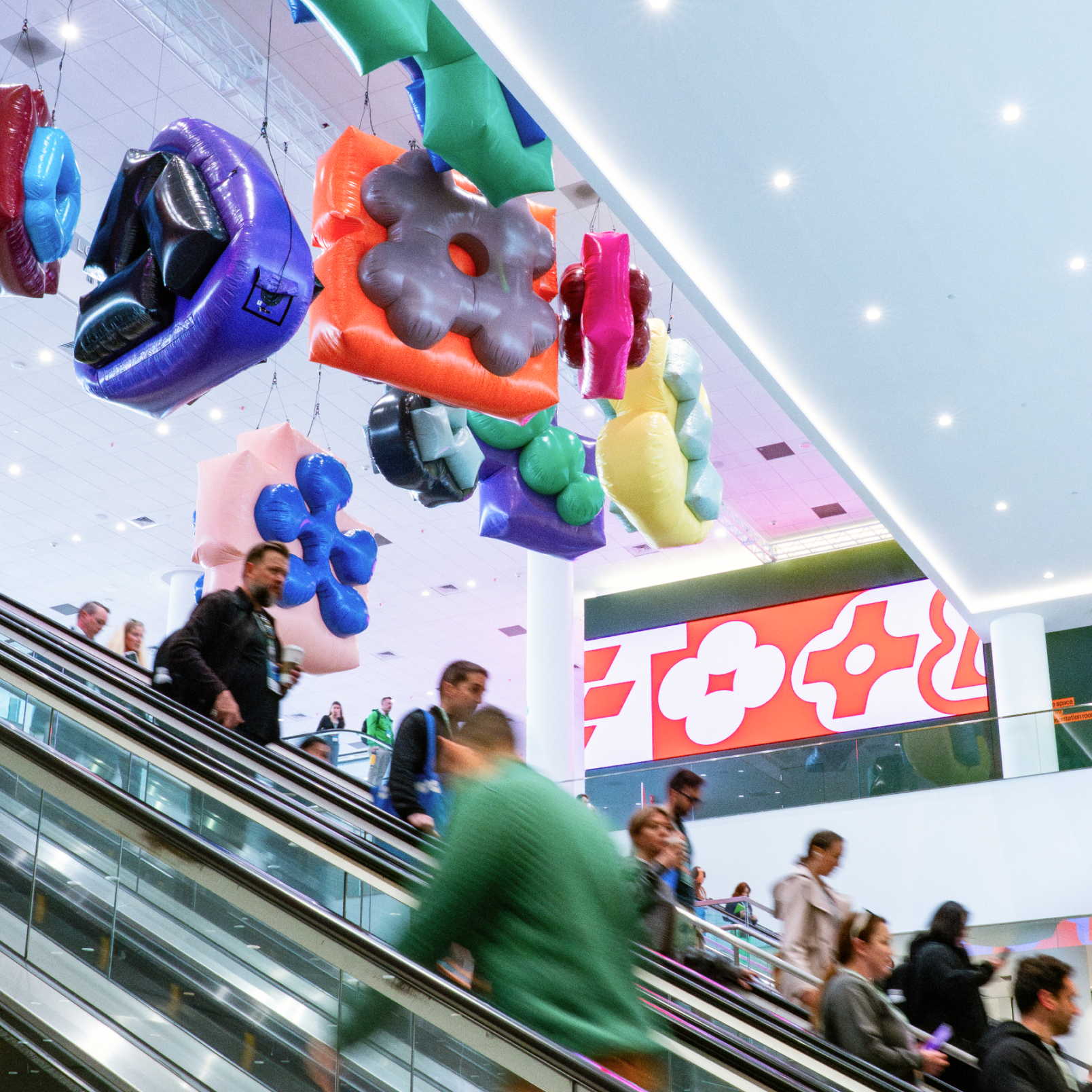 Conference attendees ride escalators below colorful inflatable shapes and large digital screen with abstract symbols.