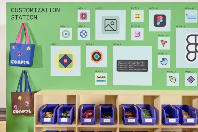 There is a green wall with text in the upper level corner saying ‘Customization Station.’ Beneath are two hanging tote bags, with three embroidered patches on each bag. To the left, there are large printed panels of the patch designs. Beneath, there is a row of blue bins with patches inside.