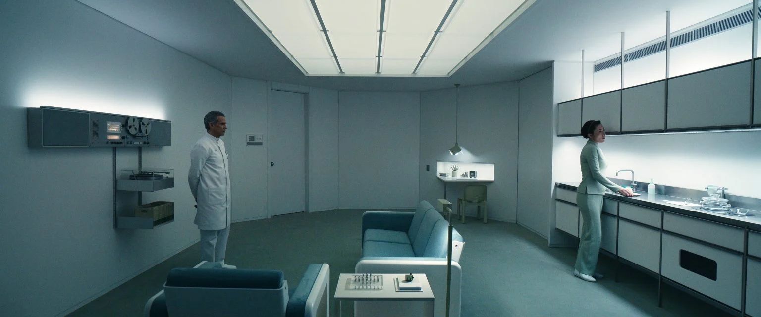 A minimalist, sterile room from Severance shows a man in white standing near a Dieter Rams audio system while a woman works at a sleek kitchen counter under bright ceiling lights.