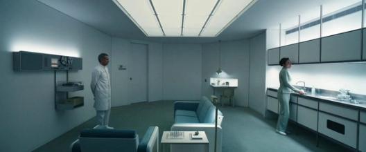A minimalist, sterile room from Severance shows a man in white standing near a Dieter Rams audio system while a woman works at a sleek kitchen counter under bright ceiling lights.
