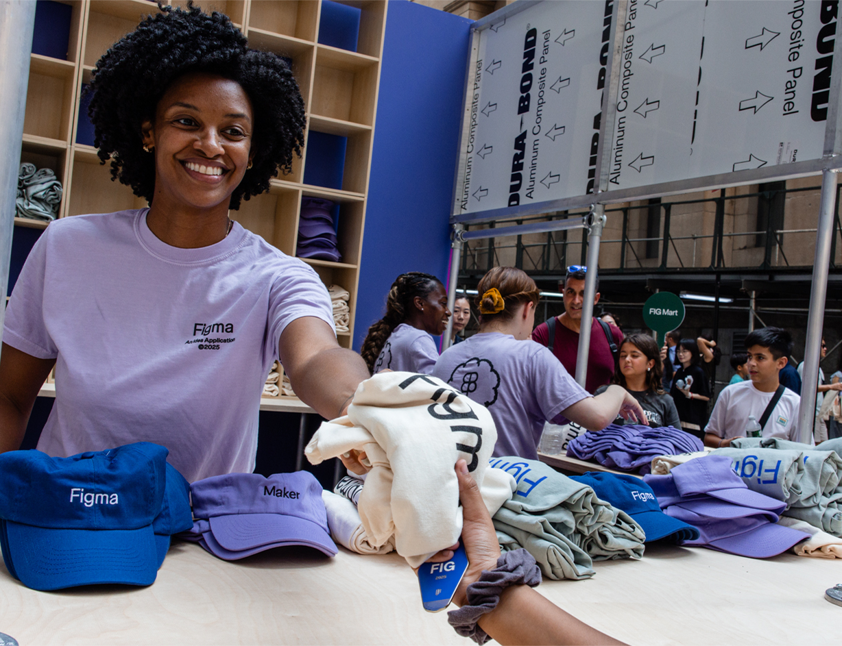 A woman hands a shirt to an attendee at a merch booth stocked with hats and apparel.