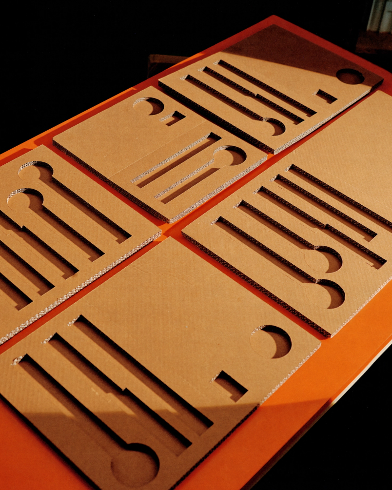 Cardboard sheets with precisely cut utensil-shaped slots arranged on orange surface under angled warm light.