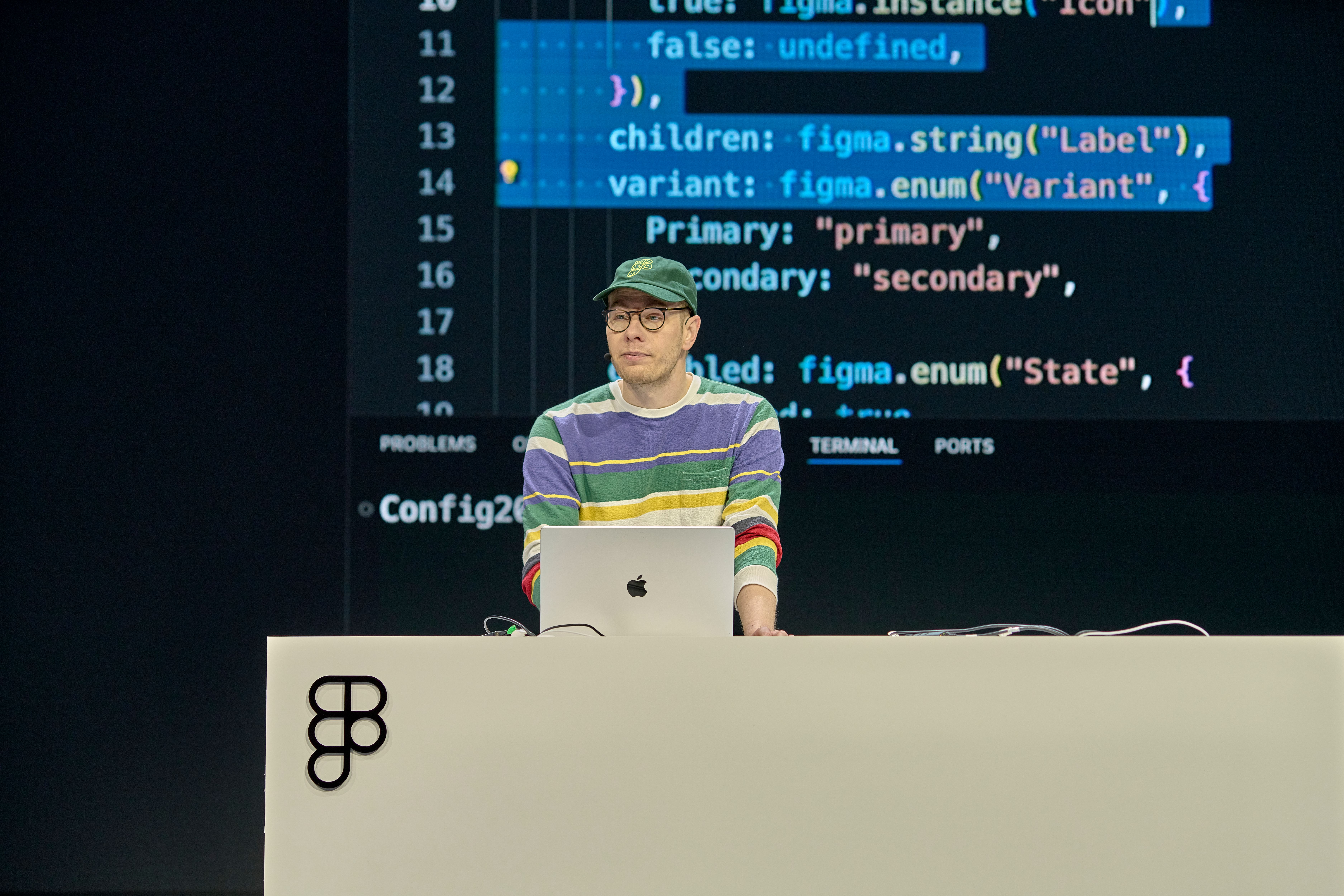 A speaker wearing a colorful striped sweater and a green cap stands behind a podium, speaking on stage. The background displays programming code projected on a large screen.