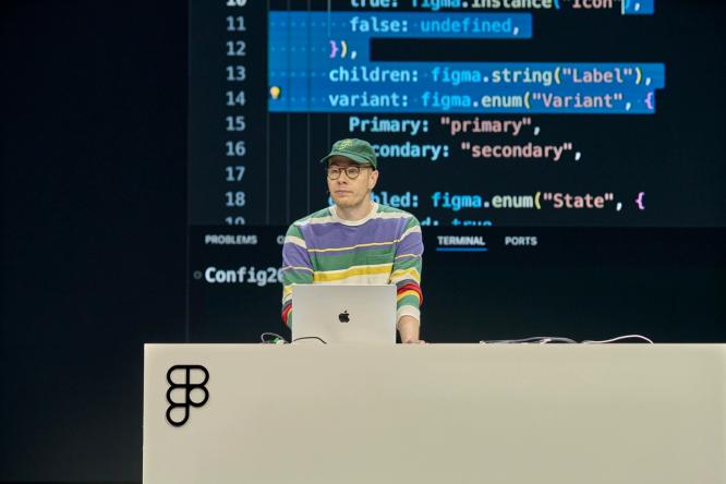 A speaker wearing a colorful striped sweater and a green cap stands behind a podium, speaking on stage. The background displays programming code projected on a large screen.