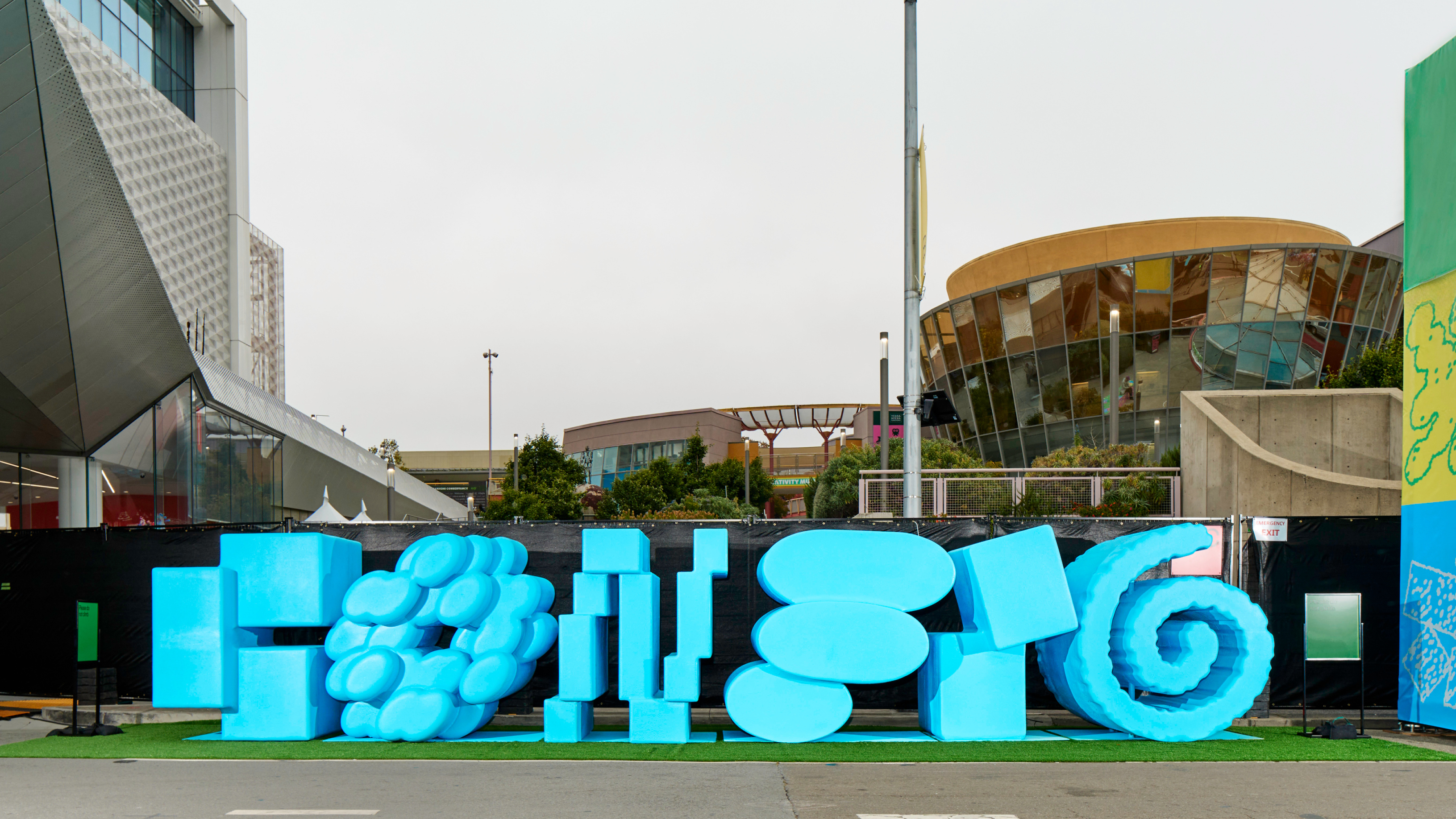 A large bright blue sculptural sign in varied abstract shapes sits on artificial turf outside a modern conference center complex.