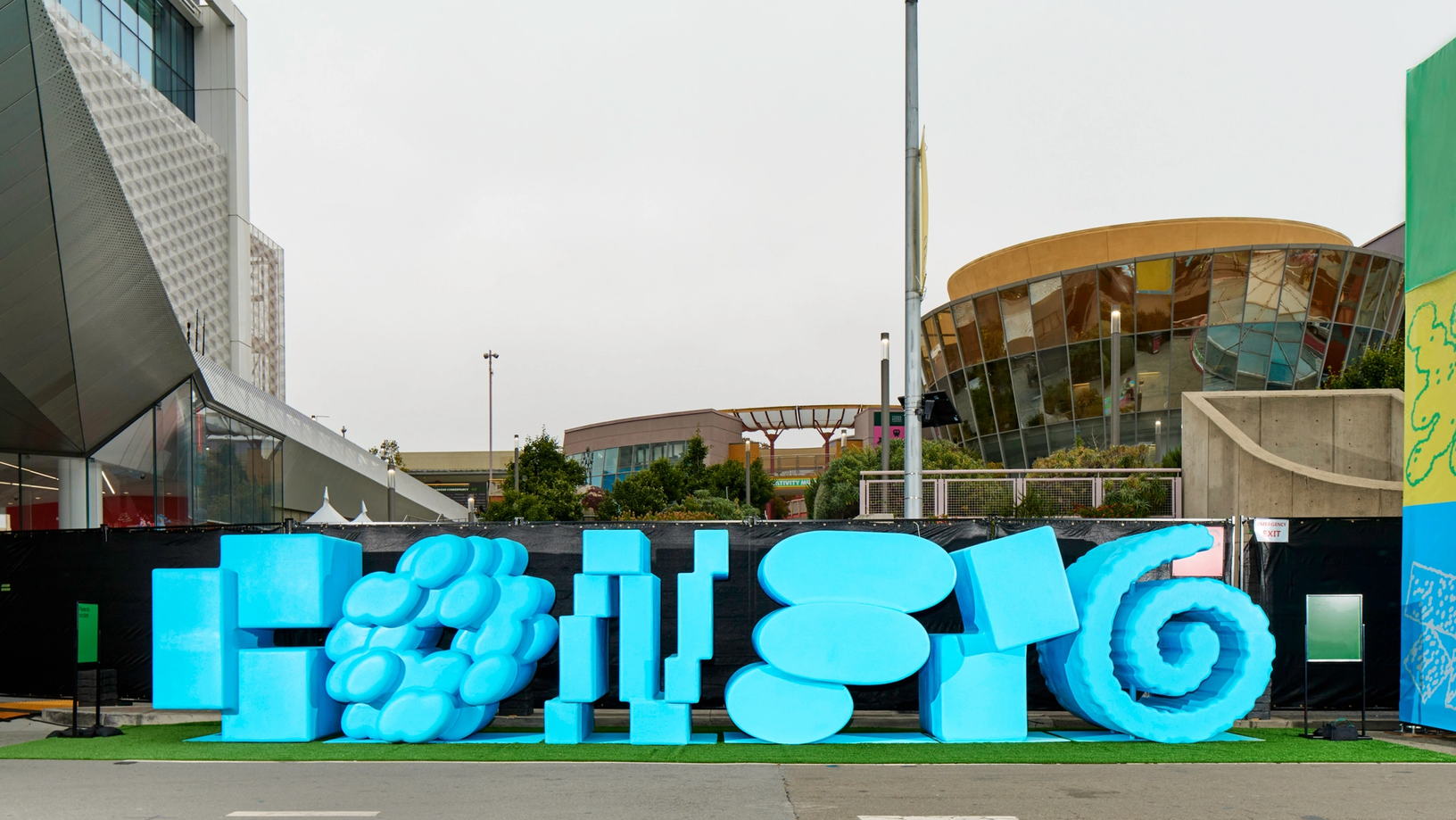 A large bright blue sculptural sign in varied abstract shapes sits on artificial turf outside a modern conference center complex.