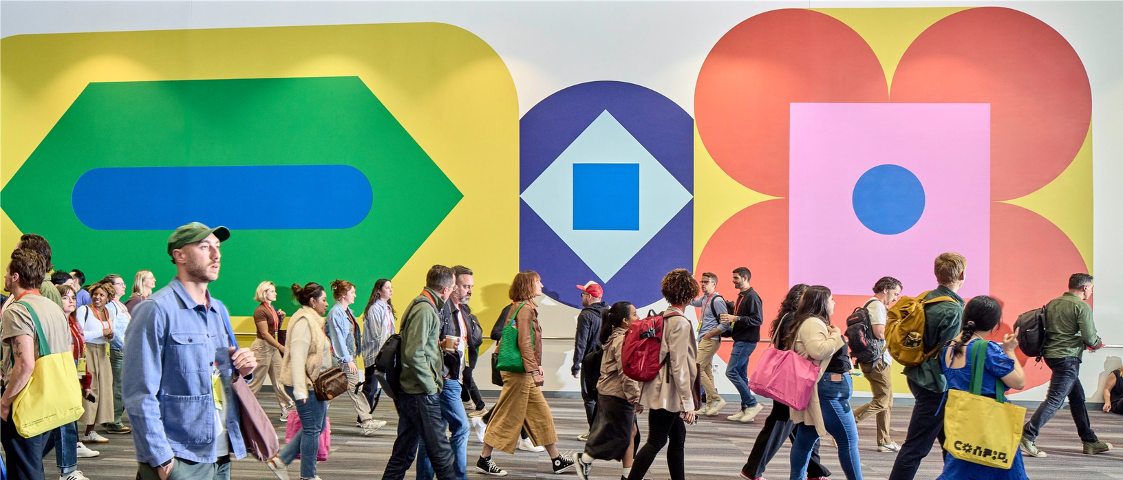 Dozens of people are walking through a large hallway, some talking together or carrying bright-colored tote bags. On the wall behind the crowd are large, 17-foot high shapes in yellow, green, blue, pink, and orange. There are three main shapes, each with nested circles, diamonds, squares, or pill shapes inside.