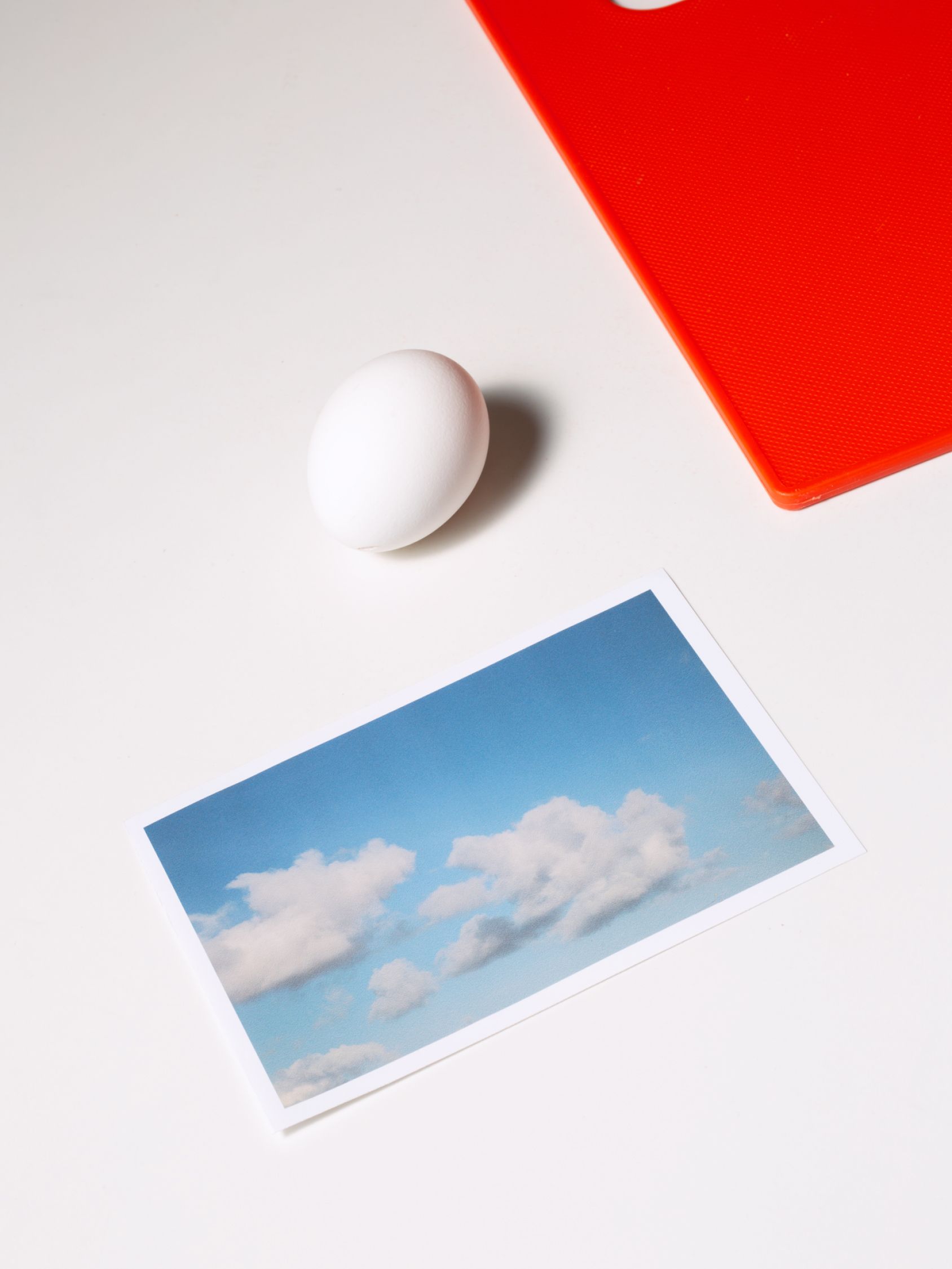 White egg, red cutting board, and a printed photo of clouds arranged on a clean white surface.