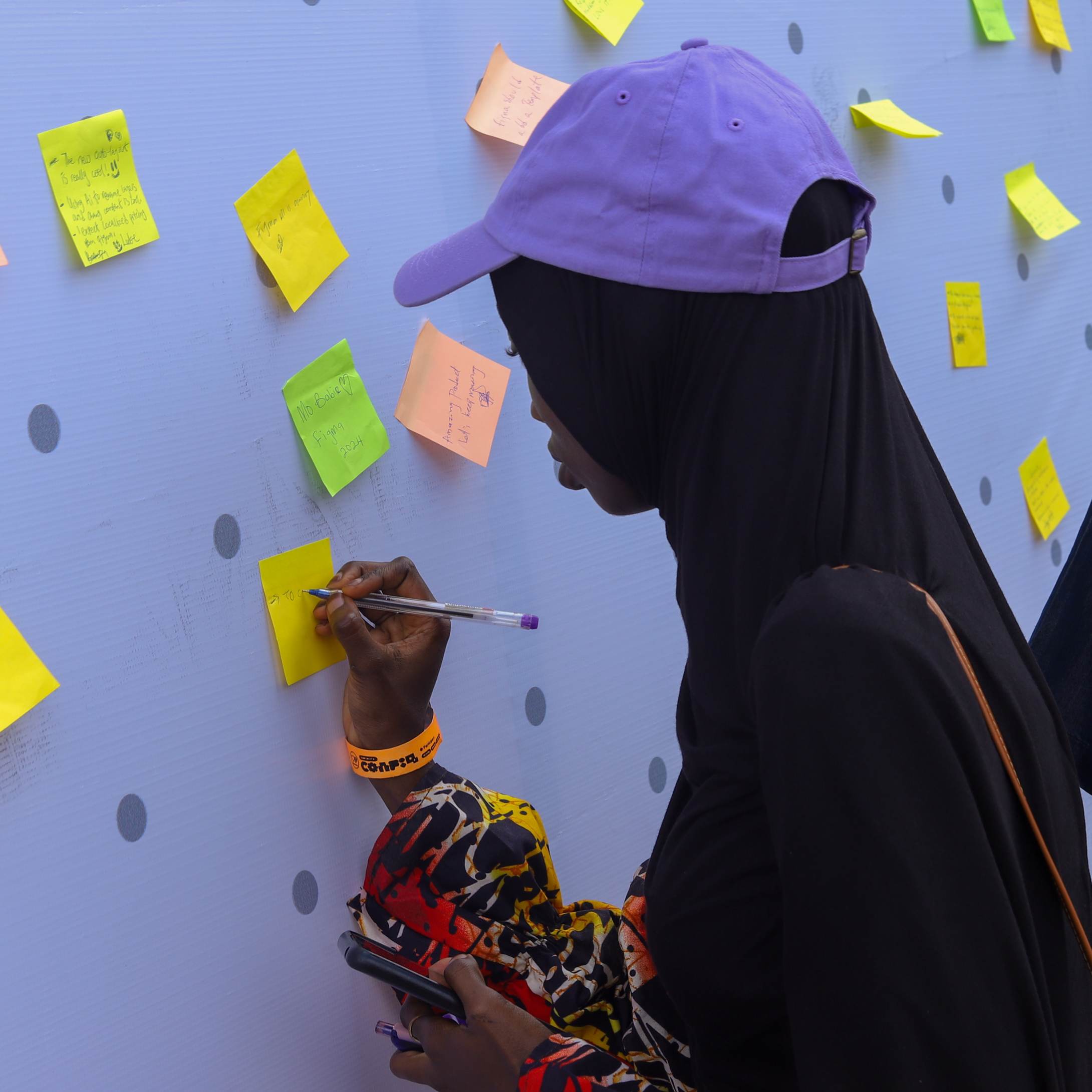 A person wearing a purple cap and colorful attire writes on a yellow sticky note attached to a whiteboard covered with multi-colored sticky notes. The scene is part of an interactive activity.