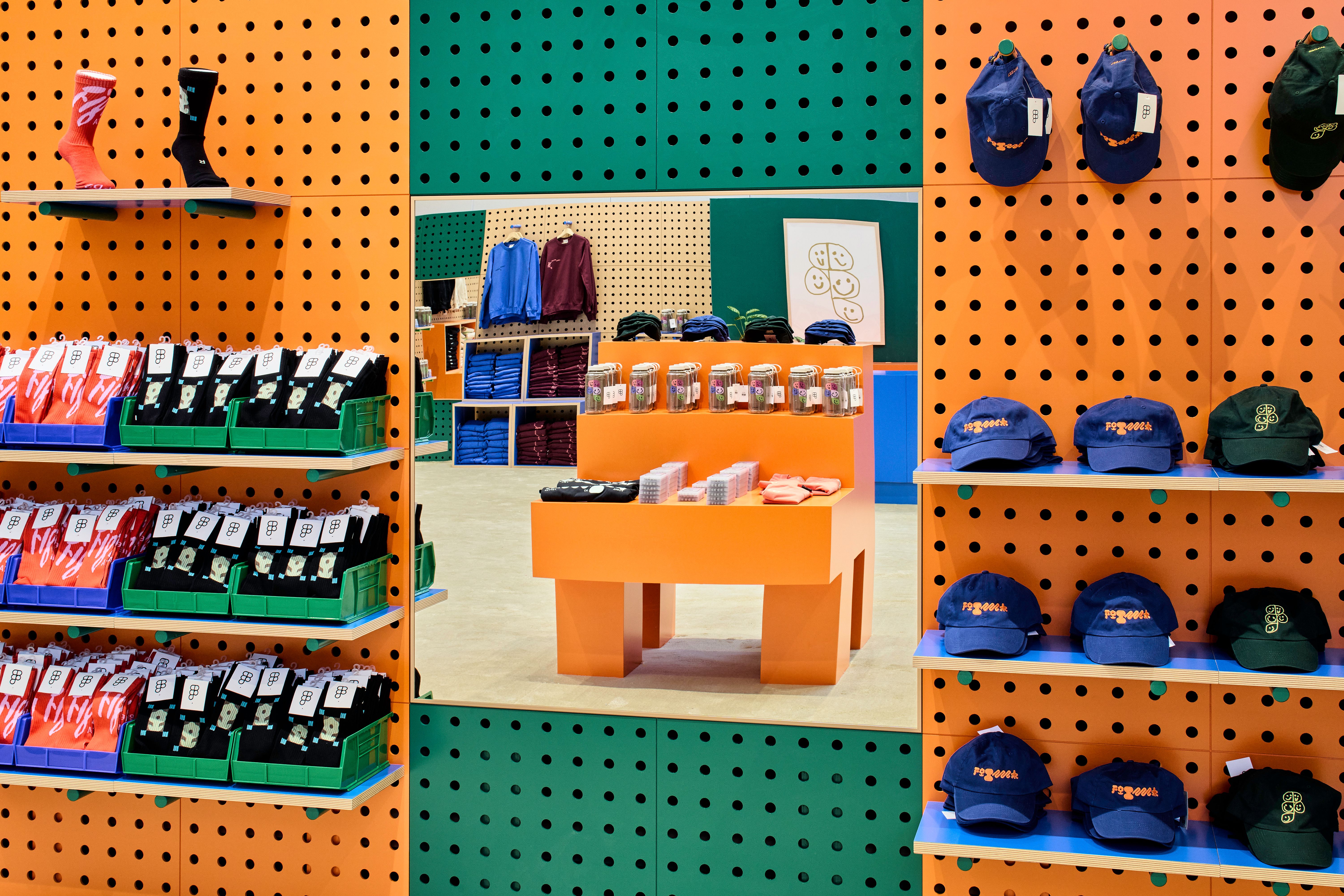 This image focuses on a orange and green wall in the Figma store, where different socks and hats are displayed on racks. In the middle of the socks and hats is a mirror, which reflects an orange table in the center of the store. 