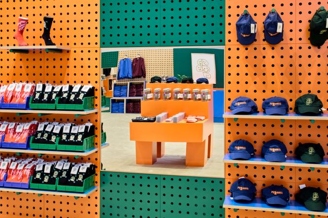This image focuses on a orange and green wall in the Figma store, where different socks and hats are displayed on racks. In the middle of the socks and hats is a mirror, which reflects an orange table in the center of the store.