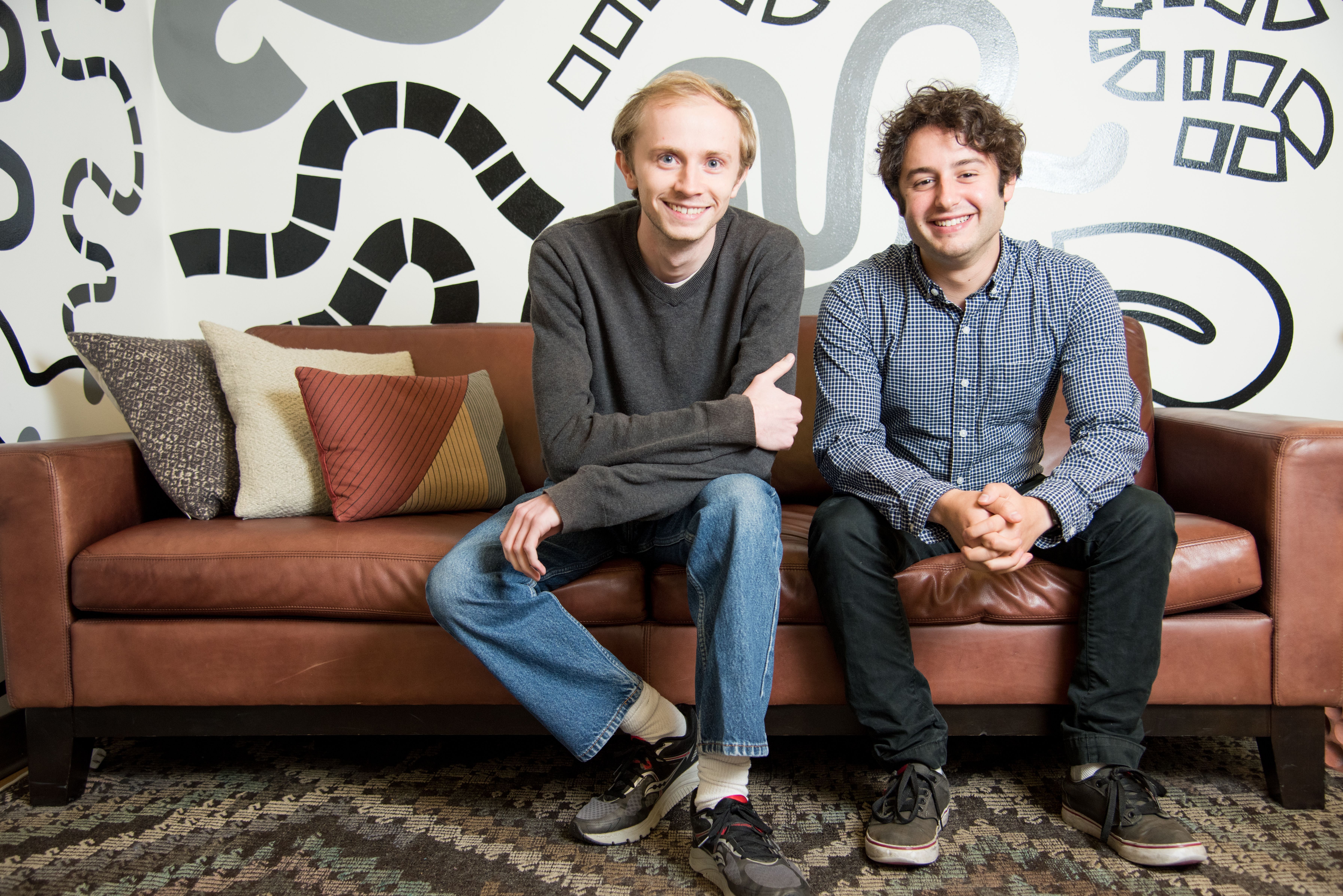 Two men sit smiling on a brown leather couch