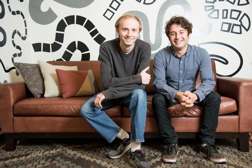 Two men sit smiling on a brown leather couch