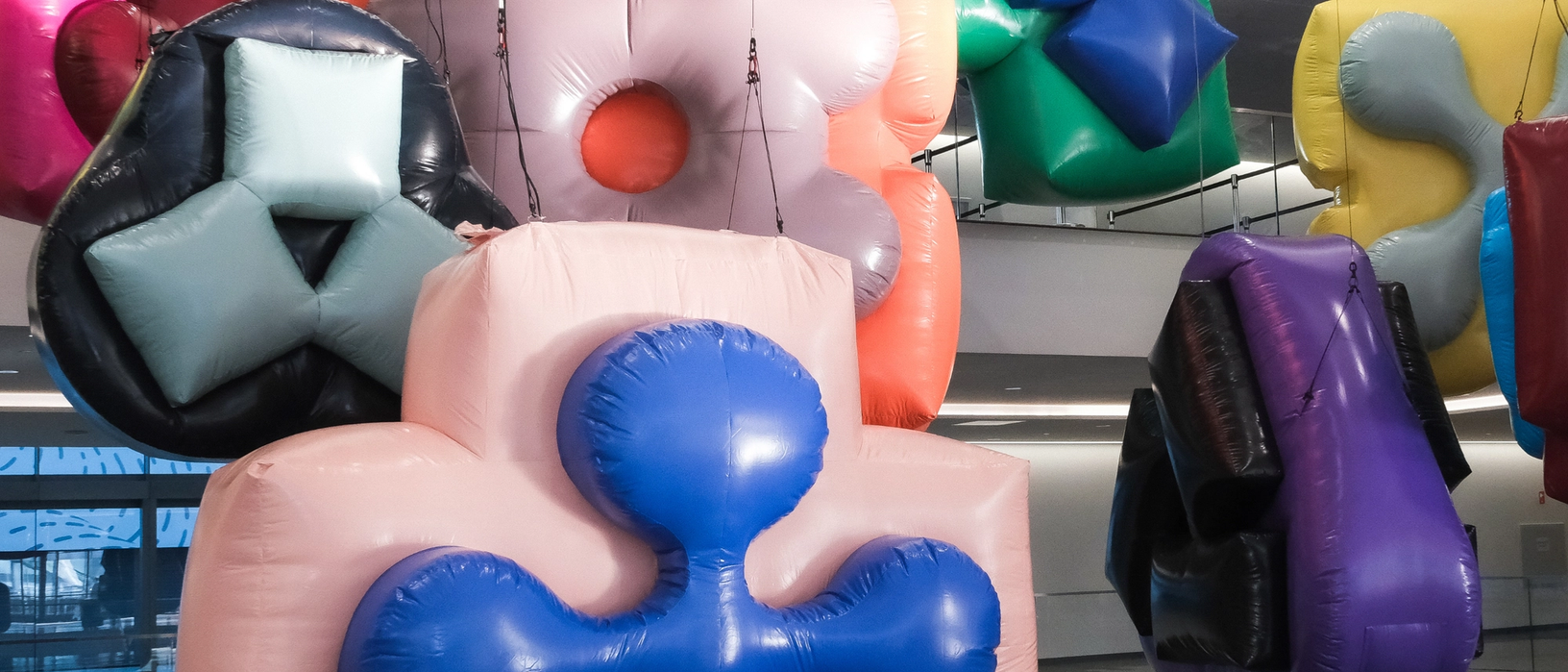 Inflatable sculptures in various abstract shapes and colors hang from a ceiling in bright indoor light